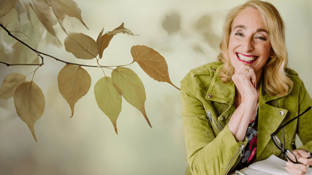 Eine lächelnde Frau mit blondem Haar sitzt vor einem Notizbuch und hält eine Brille, umgeben von grünen und braunen Blättern im Hintergrund.